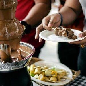 Hands dipping fruit into a chocolate fountain at a party.
