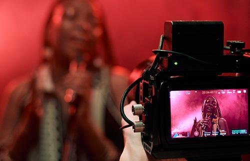 Camera filming a DJ at a party with lighting and crowd.
